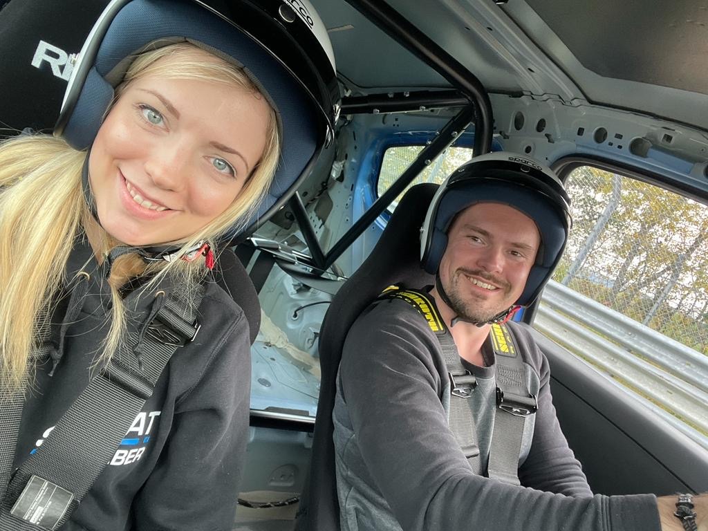 An in-car selfie of Olli, the founder of Pure Ring Rentals, and a passenger, both smiling and wearing racing helmets and full harnesses.