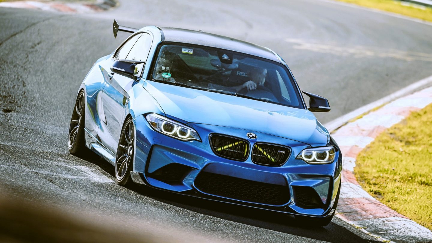 An action photograph of a blue BMW M2 Competition with a prominent rear wing driving fast on the Nürburgring circuit during Touristenfahrten, part of our rental fleet.