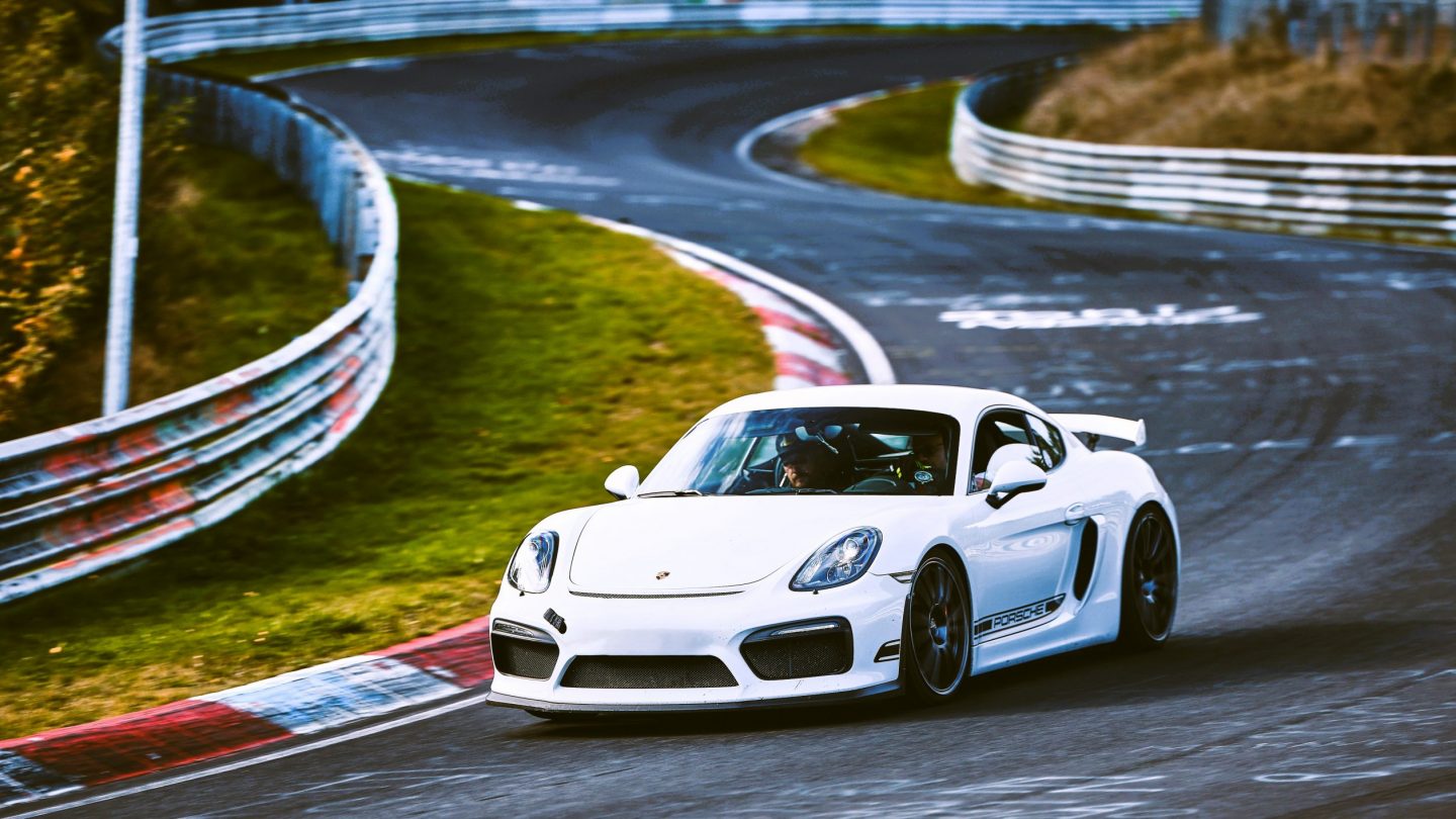 A front-quarter action photograph of a white Porsche Cayman GT4 navigating a corner on the Nürburgring Nordschleife track, representing our high-performance rental car.